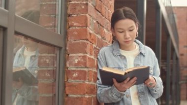 Zeki Asyalı üniversite öğrencisinin orta yavaş portresi. Kitap okuyor ve bulutlu bir günde kampüsün dışında kameraya gülümsüyor.