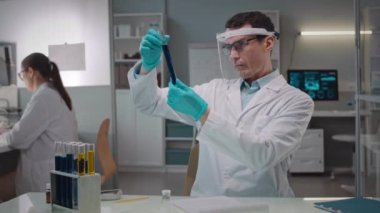 Beyaz önlüklü beyaz erkek mikrobiyolog cam test tüplerindeki sıvıların kültürünü inceliyor, bilimsel laboratuvarda asistanla birlikte çalışıyor.
