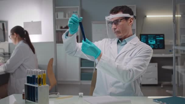 白种人专业男性微生物学家戴防护面罩和白种人实验室衣检查玻璃管中液体培养的摄像镜头 与助手一起在科学实验室工作 — 图库视频影像