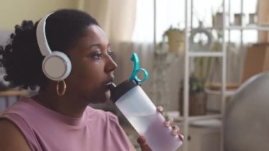 Enerjik, genç, kıvrımlı, siyah bir kadının göğsü kablosuz kulaklıkla müzik dinlerken ev çalışmasından sonra dinlenirken içme suyu içiyor.