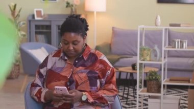 Genç, iri yarı, Afro-Amerikan bir kadın akıllı telefondan arıyor. Parlak, modern oturma odasında sandalyede oturuyor.
