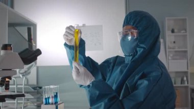 Laboratuvarda araştırma yaparken cam test tüplerindeki sıvıları inceleyen koruyucu giysili erkek biyokimyacının belden yukarı görüntüsü.