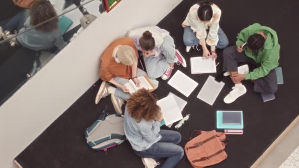 五个不同的新生坐在地板上 手里拿着大学礼堂里的书 准备一起做项目的慢镜头顶部 — 图库视频影像