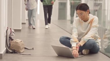 Asyalı kız öğrencilerin yavaşlaması üniversite koridorunda yerde otururken dizüstü bilgisayarda çalışıyor.