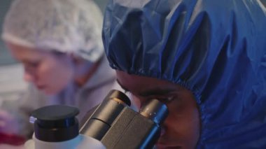 Modern araştırma laboratuarında mikroskop ile materyalleri inceleyen genç çift ırklı erkek kimyagerin ve bayan arkadaşının orta boy yakın çekimi.