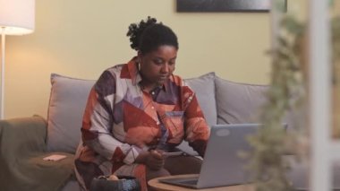 Genç ve cüsseli Afrikalı bir kadın evde alışveriş yaparken kredi kartı ve laptop kullanıyor oturma odasındaki yumuşak kanepede oturuyor.