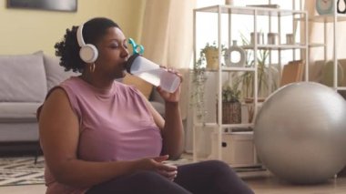 Genç, kıvrımlı, siyah bir kadının evde vücut geliştirme eğitiminden sonra dinlenirken kablosuz kulaklıkla müzik dinlediği orta boy bir fotoğraf.