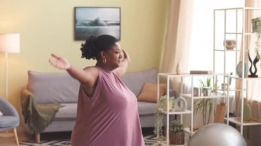 Genç kıvrımlı, aktif giyimli siyahi kadın evde yoga yaparken ayağa kalkıyor.