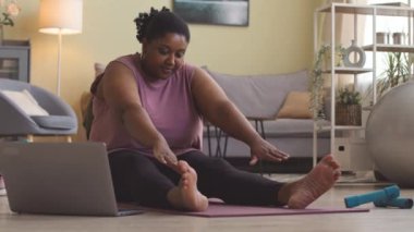 Genç ve cüsseli Afrikalı bir kadın laptopta video dersi izlerken oturma odasında paspas üzerinde esneme egzersizleri yapıyor.