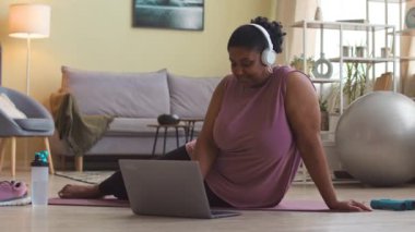 Genç ve cüsseli, kablosuz kulaklıklı ve aktif giysili bir kadın evdeki oturma odasında antrenman yaparken dizüstü bilgisayar başında egzersiz yapıyor.