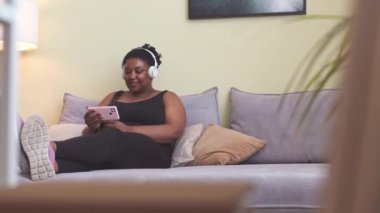 Neşeli, genç, iri yarı, kablosuz kulaklıklı Afro-Amerikan kadın. Oturma odasındaki kanepede oturmuş akıllı telefondan komik videolar izlerken gülüyorlar.