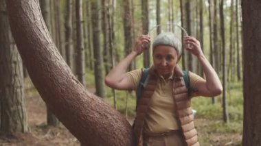 Ormanda ağaç gövdesine yaslanırken kameraya gülümseyen kablosuz kulaklıklı Asyalı fit kadının orta boy portresi.