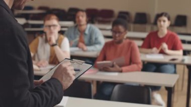 Kafkas sakallı üniversite profesörünün oditoryumdaki masalarda oturan çok kültürlü öğrencilere ders verirken eliyle not tuttuğu görüntüler.