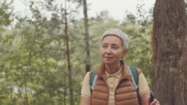 Yetişkin Asyalı bir kadının İskandinav yürüyüşü sırasında çevreye bakınıp orman doğasının tadını çıkardığı belden yukarı çekimler.