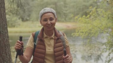 Yaşlı Asya 'lı turistin elinde yürüyüş direğiyle ormanda yürürken kameraya konuşan orta boy fotoğrafı.