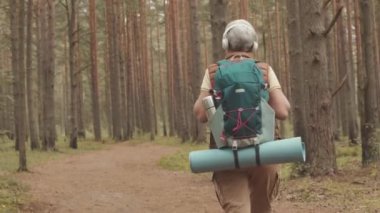 Yaşlı kadın turistin kulaklıklı, sırt çantalı, gündüz vakti ormanda yürüyüş yaparken orta açıdan çekilmiş bir fotoğrafı.