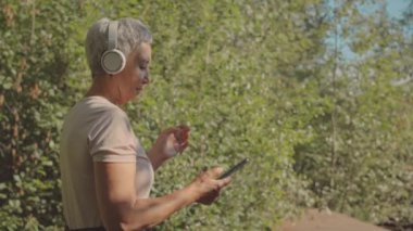 Aktif, olgun Asyalı bir kadının kablosuz kulaklıklarıyla gündüz vakti ormanda koşarken akıllı telefonuyla müzik açtığı orta boy bir fotoğraf.
