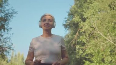 Güneşli bir günde parkta koşan, kablosuz kulaklıklı, aktif, yaşlı Asyalı bir kadının orta boy fotoğrafı.