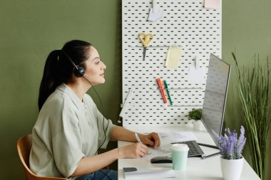 İş yerindeki kulaklıklı genç iş kadını bilgisayar monitörünün önünde oturuyor ve internette konuşuyor.