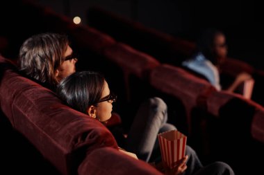 Genç bir çiftin sinemada rahat koltuklarda gözlük takıp 3D film izlemesi.