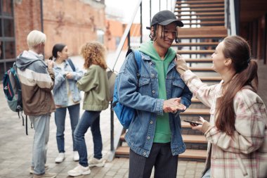 Kot ceketli ve şapkalı mutlu adam beyaz kız arkadaşıyla konuşuyor elini arkadaşlarına karşı omzunda tutuyor.