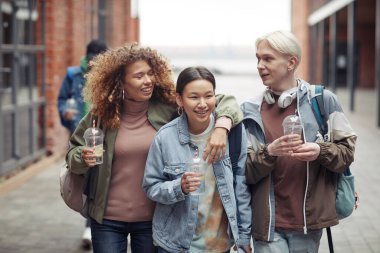 Sıradan giyimli, plastik bardakta içki içen ve üniversiteden eve dönerken komik şeyleri tartışan üç genç arkadaş.
