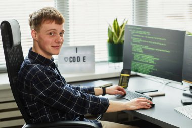 İş yerinde bilgisayar monitörü ve yazım kodlarıyla otururken kameraya bakan genç programcının portresi