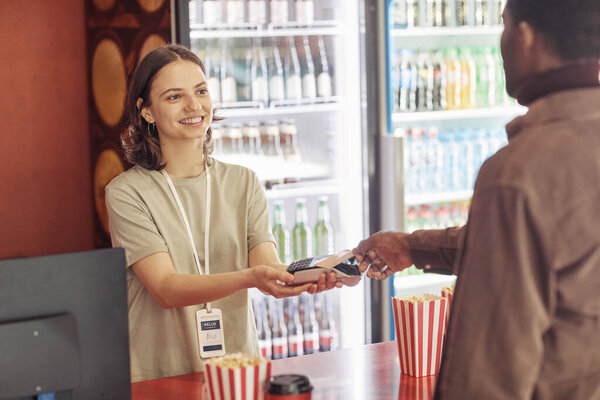 Young smiling seller holding terminal while customer paying for popcorn with credit card in the shop