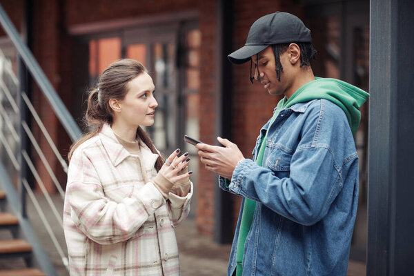 Side view of youthful multicultural couple in casualwear having chat while African American guy texting in mobile phone in the street