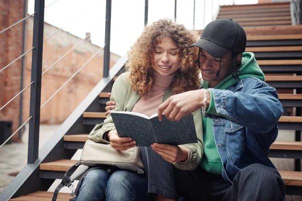 stock image Two adolescent students sitting on staircase and reading book or looking through lecture notes while happy guy embracing his girlfriend