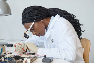 Laboratuvarda elektronik parçaları inceleyen siyah kadın mühendisin yan görünüm portresi.