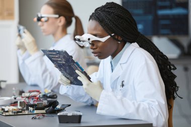 Büyüteç takan ve kalite kontrol laboratuarında elektronik parçaları inceleyen siyah bir kadının portresi.