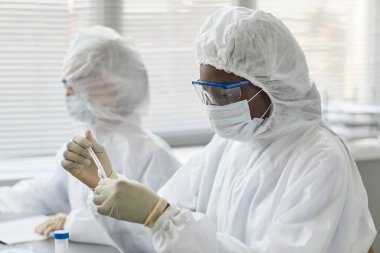 Tıbbi laboratuvarda tehlikeli numunelerle araştırma yaparken koruyucu giysiler giyen genç kadın bilim adamının portresi.