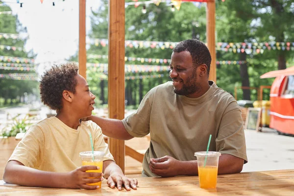 Happy dad talking and laughing with his son while they drinking cold juice at table in outdoor cafe
