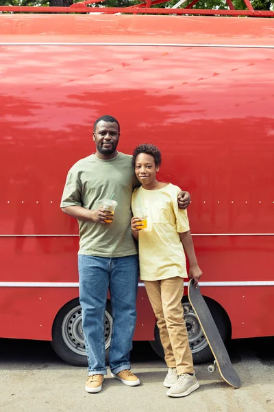 Portrait of African dad embracing his son and looking at camera while they refreshing with cold drinks after walking standing against red van