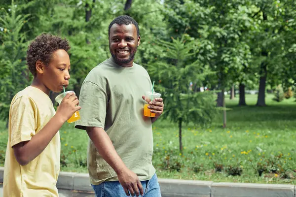 Happy African dad drinking cold juice and having a stroll with his son in the park during summer vacation