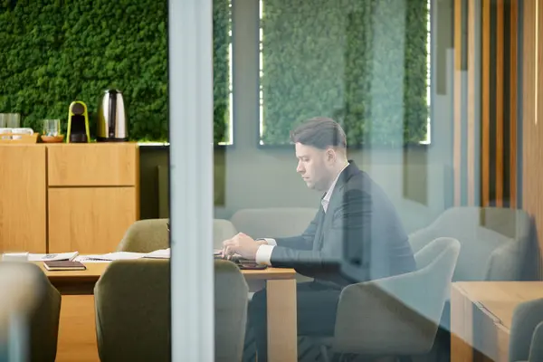 Side view portrait of businessman using laptop in conference room with green eco design behind glass wall, copy space