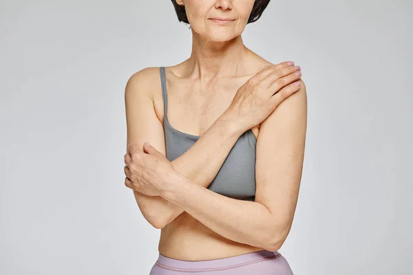 Minimal cropped shot of mature woman wearing neutral underwear against grey background, covering