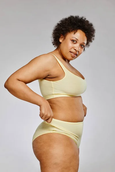 Vertical portrait of black woman wearing underwear and looking at camera against grey background, body positivity concept