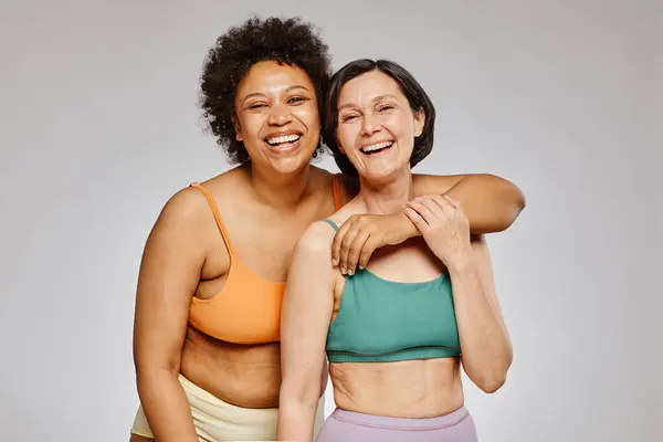 Minimal waist up portrait of two real women wearing underwear and laughing happily against grey background