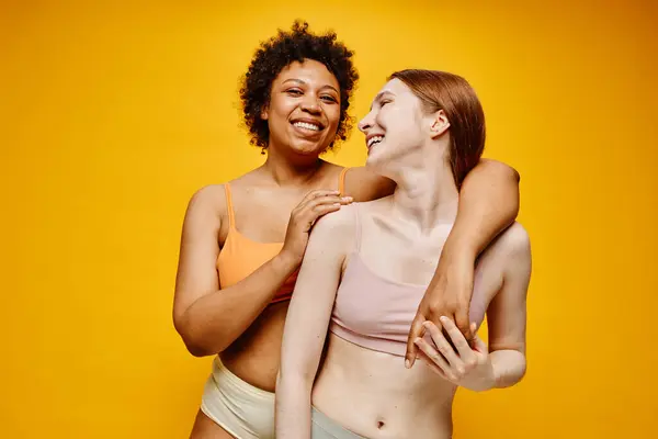 Waist up portrait of two carefree young women dark skin and fair skin embracing while standing against vibrant yellow background in underwear