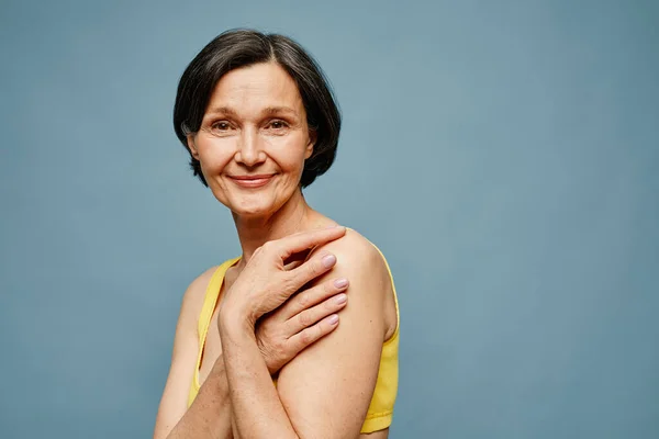 Candid portrait of mature woman smiling at camera while posing elegantly against pastel blue background, copy space