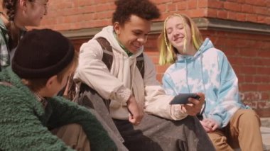 Güneşli bir günde akıllı telefondan video izleyen etnik çeşitliliğe sahip genç arkadaşların yavaşlaması.