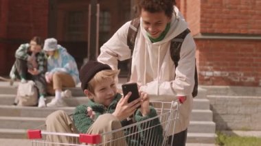 Beyaz tenli, alışveriş arabasında oturan ve yazın dışarıda takılan melez arkadaşına akıllı telefondan bir şey gösteren bir çocuk.