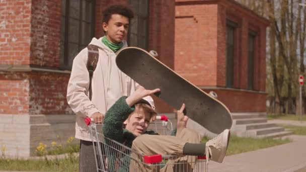 夏に屋外で会う若いケアフリーの多様な十代の企業の中間スローモ スケートボード付きのショッピングカートに座っている少年の一人 — ストック動画