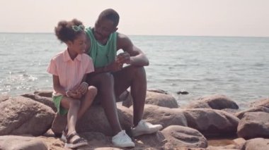 Afrika kökenli Amerikalı baba ve küçük kızı yazın kumsalda taşlarda otururken deniz kabuğuna bakıp konuşuyorlar.