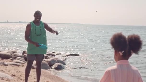 陽気な黒人の男と彼の小さな娘は晴れた日に砂浜でフリスビーと遊んでいます — ストック動画