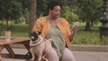 Gündüz vakti yeşil parktaki ahşap masada otururken akıllı telefon kullanan orta yaşlı siyahi kadın.