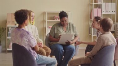 Çok ırklı gençlerden oluşan yavaş bir grup parlak modern ofiste oturup siyahi kadın psikoloji uzmanıyla terapi seansı yapıyorlar.