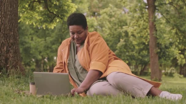 Parkın Yeşil Çimlerinde Otururken Laptopta Çalışan Orta Yaşlı Siyahi Kadın — Stok video